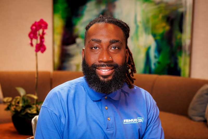 Professional headshot of a person wearing a blue Fort Bend MUD 23 polo shirt, seated indoors with decorative artwork and a flowering plant in the background.