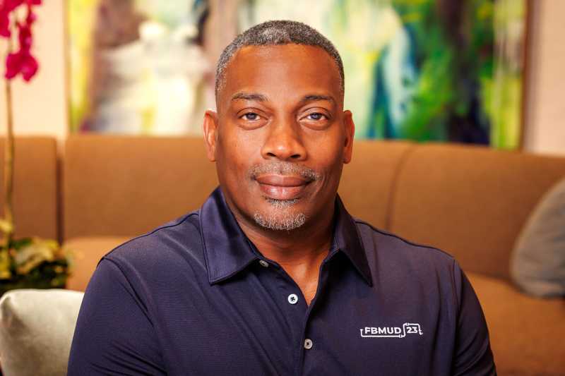 Professional headshot of a person wearing a navy Fort Bend MUD 23 polo shirt, seated indoors with abstract artwork, a flowering plant, and softly blurred background decor.