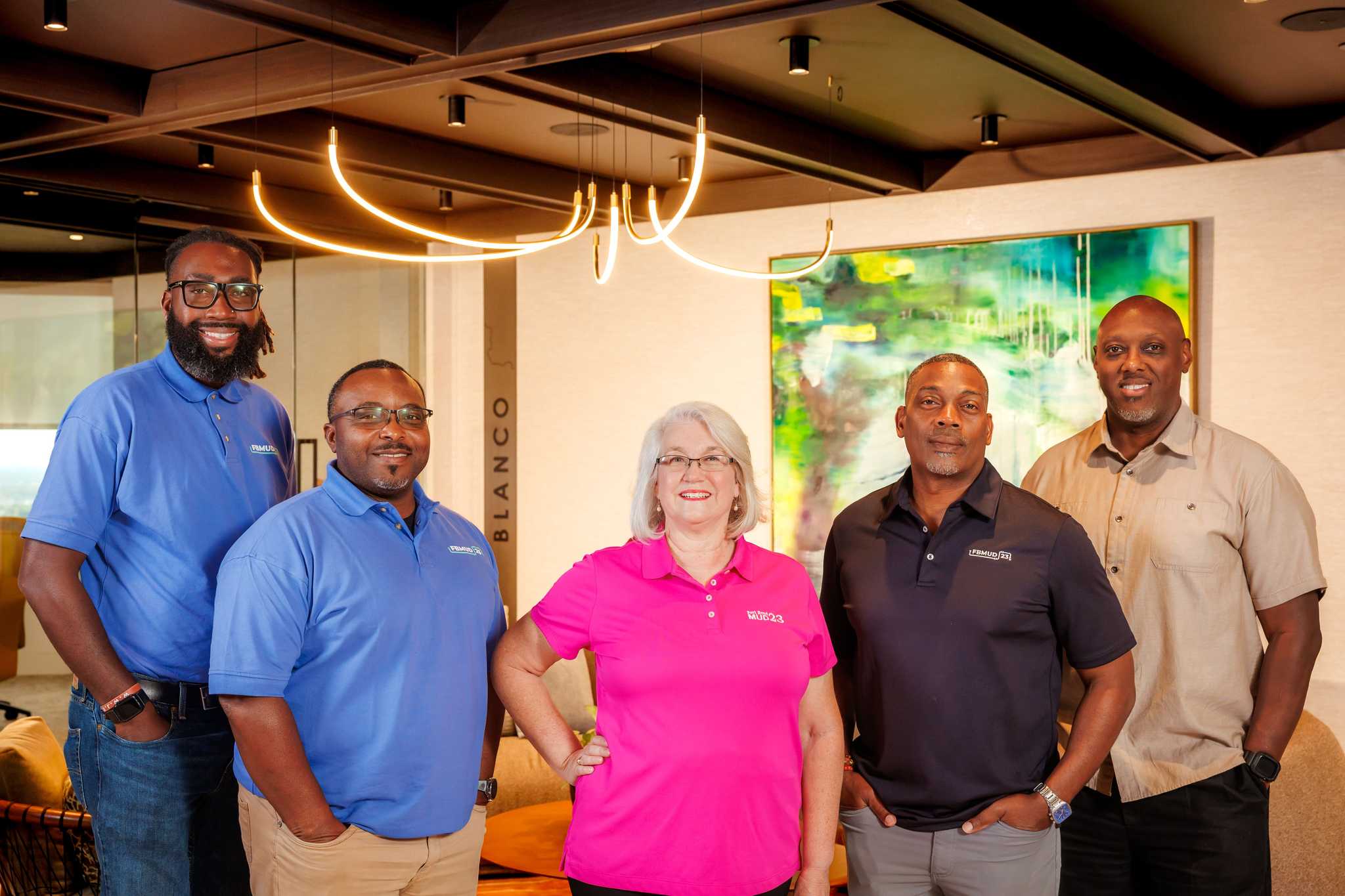Group portrait of five people wearing Fort Bend MUD 23 branded shirts, standing in a modern office lobby with decorative lighting and artwork in the background.